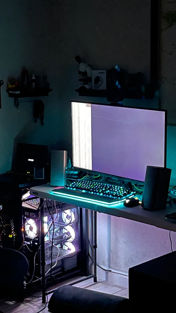 Shivam's desk at night: a liquid-cooled PC glowing under RGB, a mechanical keyboard, a monitor, and a microscope on the shelf above.